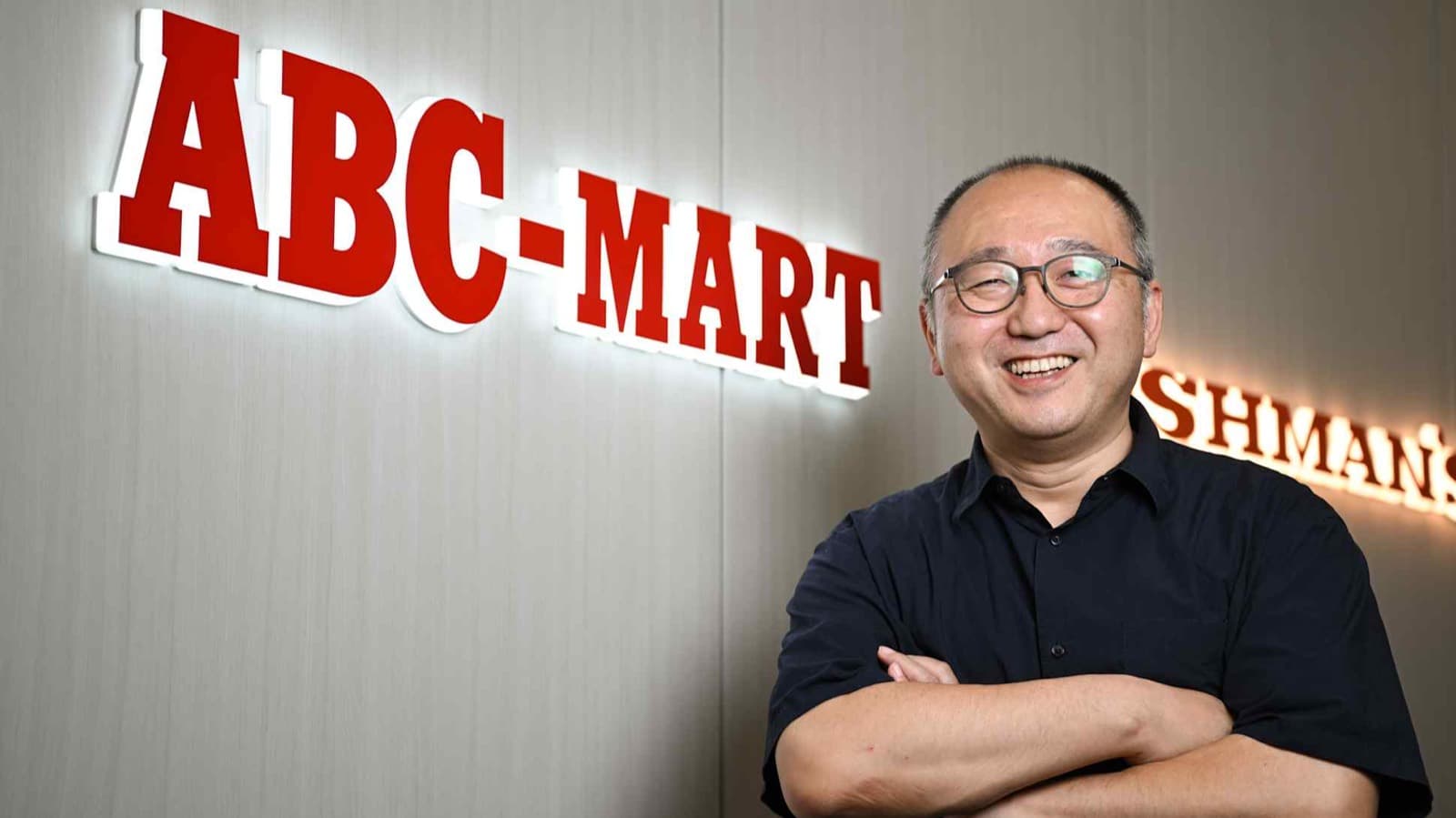 ABC-Mart president Kiichiro Hattori stands smiling beside the company's illuminated logo