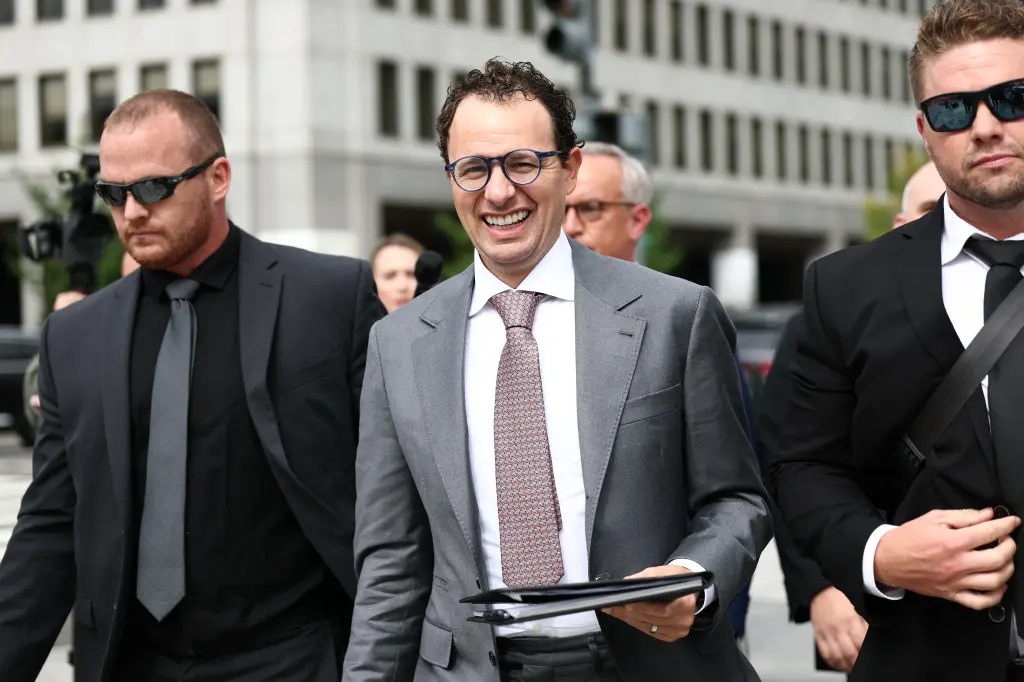 A man in glasses and a grey suit smiling while walking outdoors flanked by two security personnel in dark suits and sunglasses