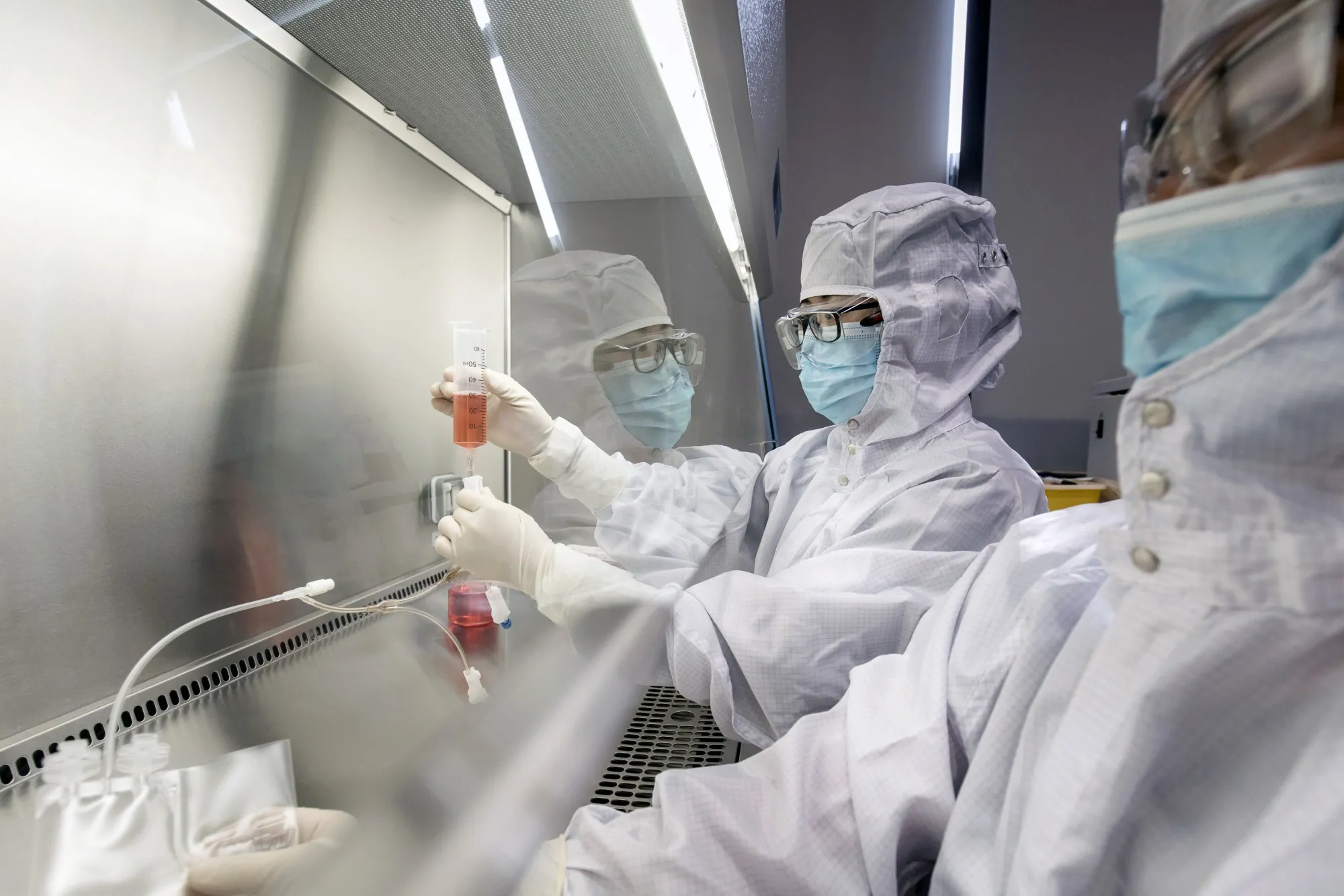 A researcher in protective cleanroom gear handling a syringe and vial inside a laboratory biosafety cabinet