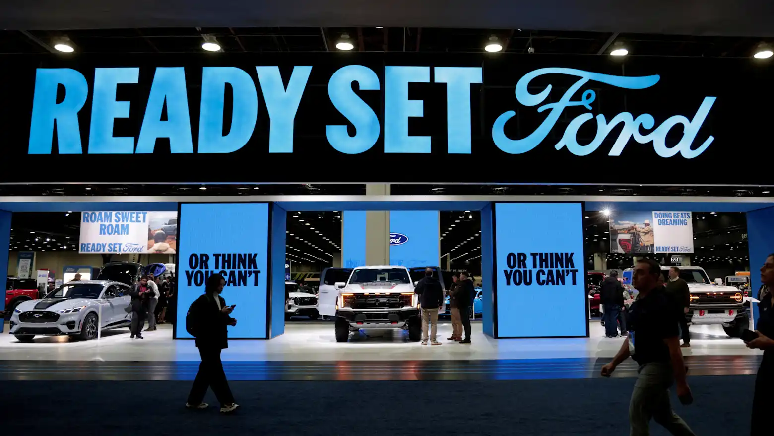 Visitors walking past a large Ford display booth with vehicles at an auto show