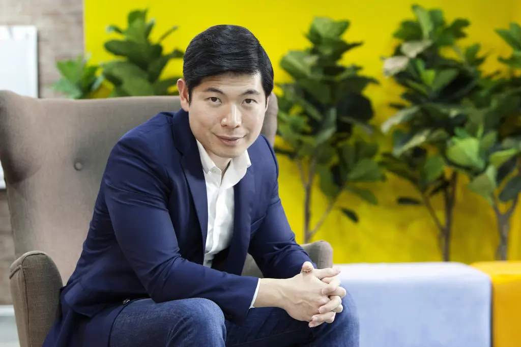 A man in a navy blazer sitting in an armchair, hands clasped, in front of a yellow wall with potted plants