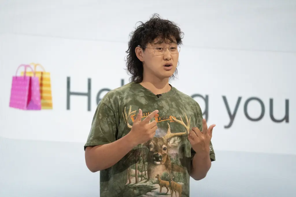 A person with curly hair and glasses speaking on stage and gesturing with both hands in front of a presentation screen