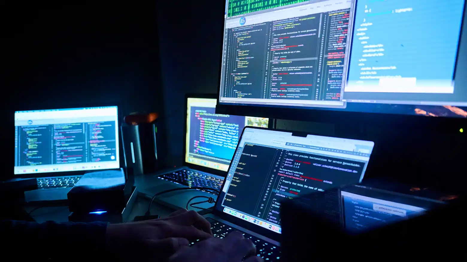 A person typing on a laptop surrounded by multiple screens displaying lines of code in a dark room