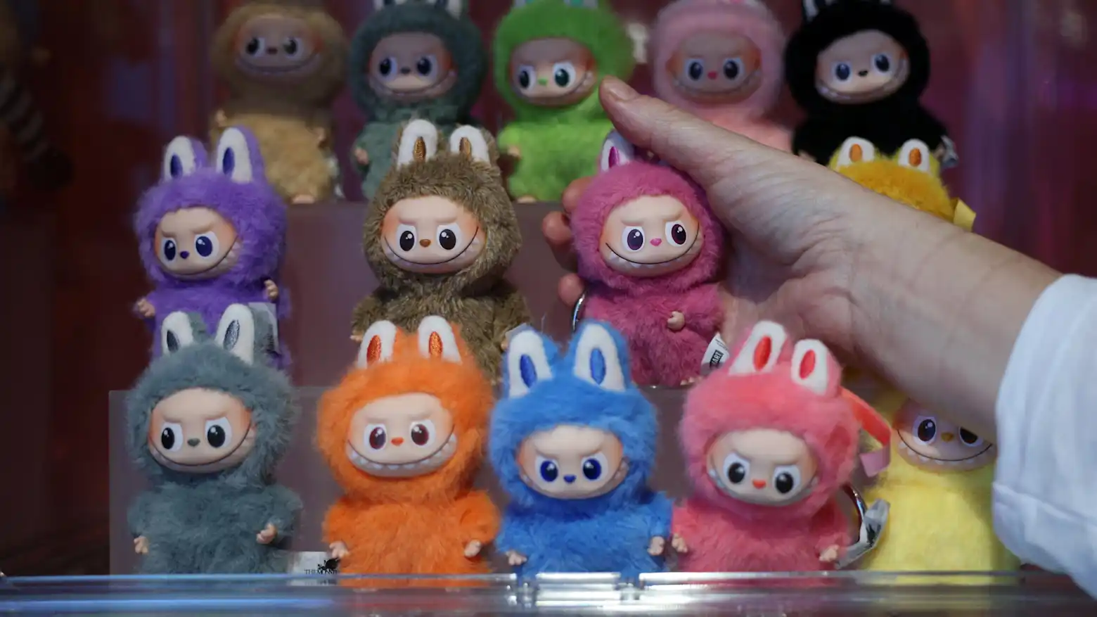 Rows of colorful furry collectible character dolls displayed on shelves, with a hand reaching toward one