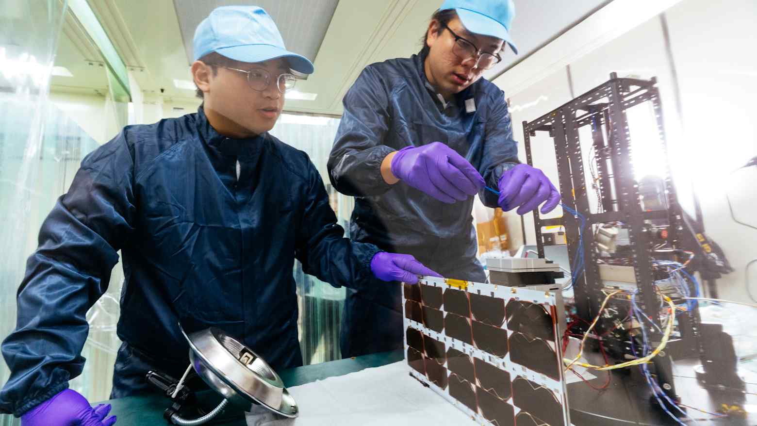 Two technicians in a cleanroom assemble a small satellite with solar panels at a workbench