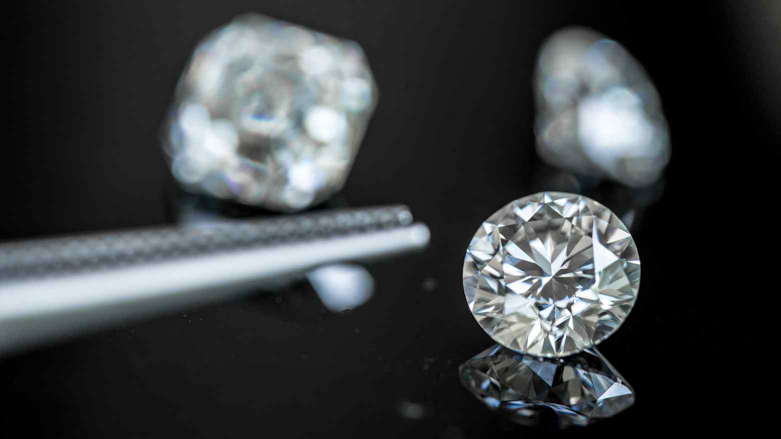 Cut and rough diamonds on a dark reflective surface, one brilliant round stone in sharp focus beside tweezers
